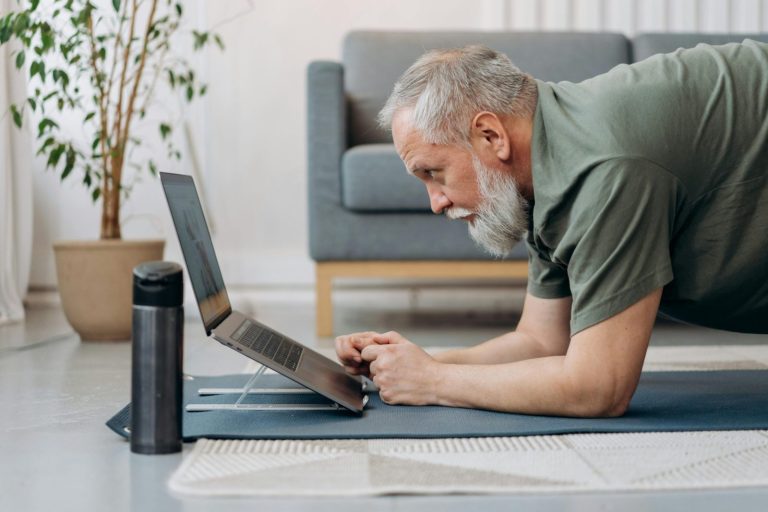 Elderly man practicing yoga on a mat while watching online instructions on a laptop.